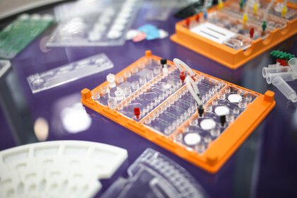 Close-up of a laboratory microplate with wells on an orange holder with blurred laboratory equipment in the background.