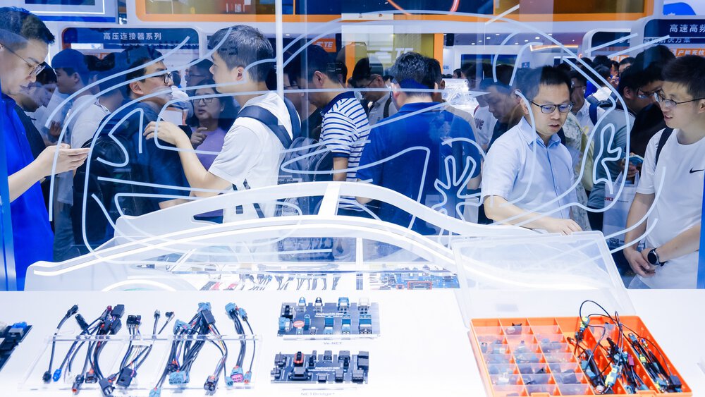 At an exhibition, electronic car components are shown behind a glass pane with the outline of a car.