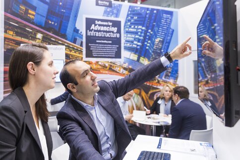 At a trade fair stand, an exhibitor demonstrates something to a visitor on a screen. In the background, three people are chatting at a small table.