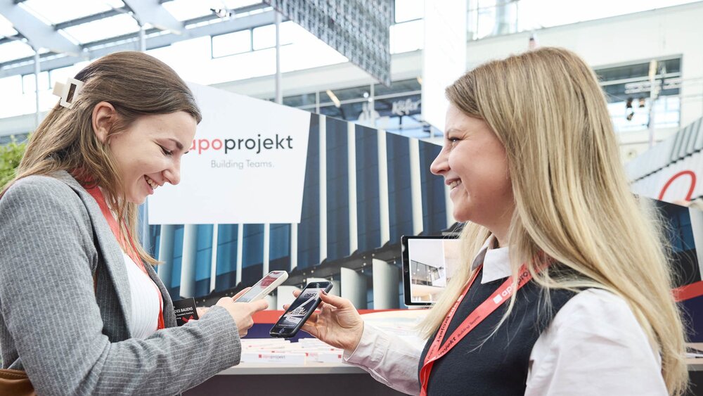 Two trade fair attendees smile as they exchange contact details by holding their smartphones together. In the background, a booth with the “apoprojekt” logo and modern architectural elements is visible.
