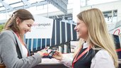 Two trade fair attendees smile as they exchange contact details by holding their smartphones together. In the background, a booth with the “apoprojekt” logo and modern architectural elements is visible.