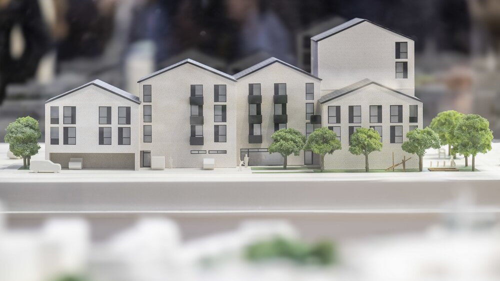 Ein maßstabsgetreues Modell eines modernen Wohnkomplexes mit mehreren Stockwerken, dunklen Fenstern und grünen Bäumen davor.