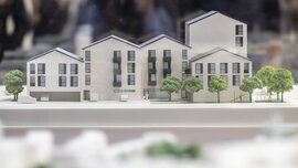 Ein maßstabsgetreues Modell eines modernen Wohnkomplexes mit mehreren Stockwerken, dunklen Fenstern und grünen Bäumen davor.