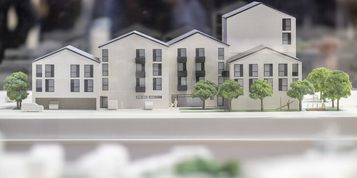Ein maßstabsgetreues Modell eines modernen Wohnkomplexes mit mehreren Stockwerken, dunklen Fenstern und grünen Bäumen davor.