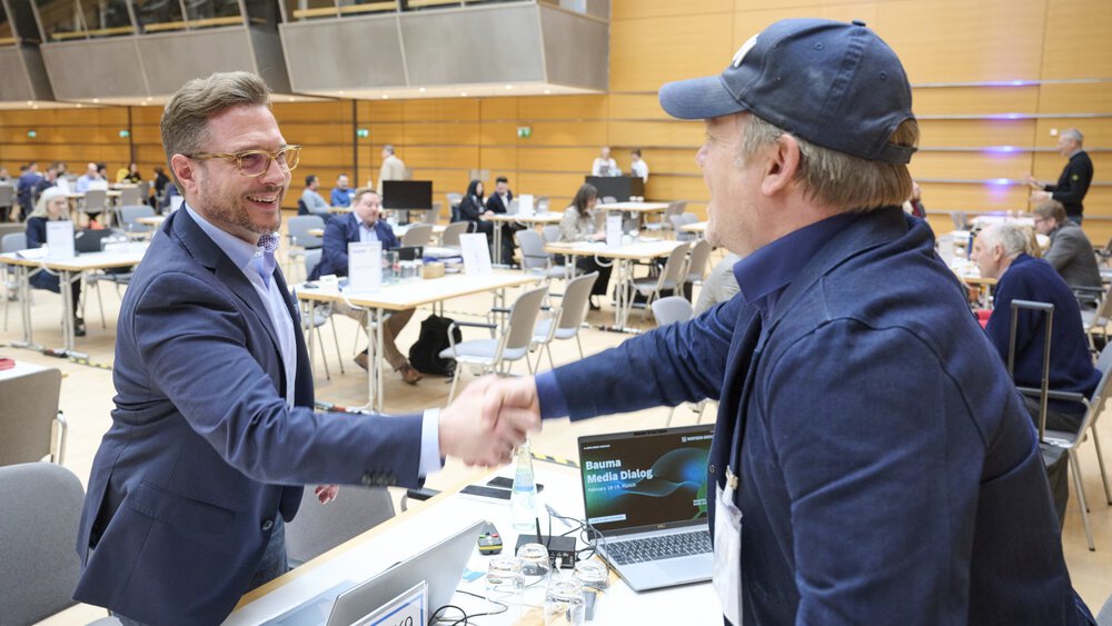 In a busy conference room, two men in business attire are shaking hands, while multiple booths and large screens in the background display presentations.