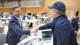 In a busy conference room, two men in business attire are shaking hands, while multiple booths and large screens in the background display presentations.