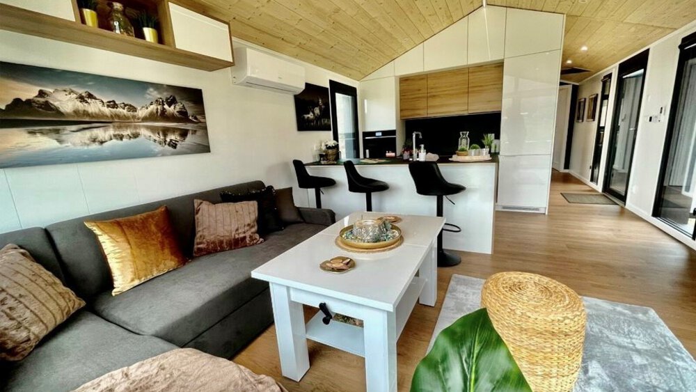 Modernes Wohnzimmer mit grauem Sofa, weißem Couchtisch, Küche mit Barhockern und Wandkunst mit einer Bergszene.