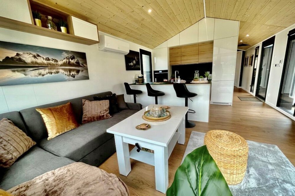 Modernes Wohnzimmer mit grauem Sofa, weißem Couchtisch, Küche mit Barhockern und Wandkunst mit einer Bergszene.