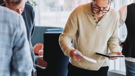 Several people are standing at a table looking at the wristwatches displayed there in black wooden boxes.