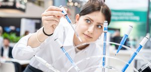 Eine Person im weißen Laborkittel, die mit einer Pipette blaue Flüssigkeit in mehrere Reagenzgläser überträgt.