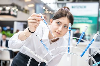 A person in a white lab coat using a pipette to transfer blue liquid into several test tubes.