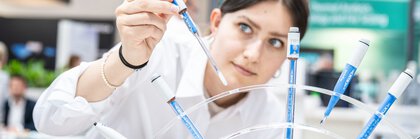A person in a white lab coat using a pipette to transfer blue liquid into several test tubes.