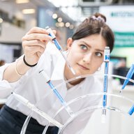 A person in a white lab coat using a pipette to transfer blue liquid into several test tubes.