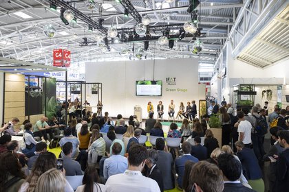 Large crowd seated and standing in front of a stage with five panelists and a screen, with „IFAT Munich Green Stage“ in the background.