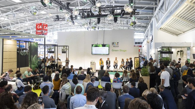 Large crowd seated and standing in front of a stage with five panelists and a screen, with „IFAT Munich Green Stage“ in the background.