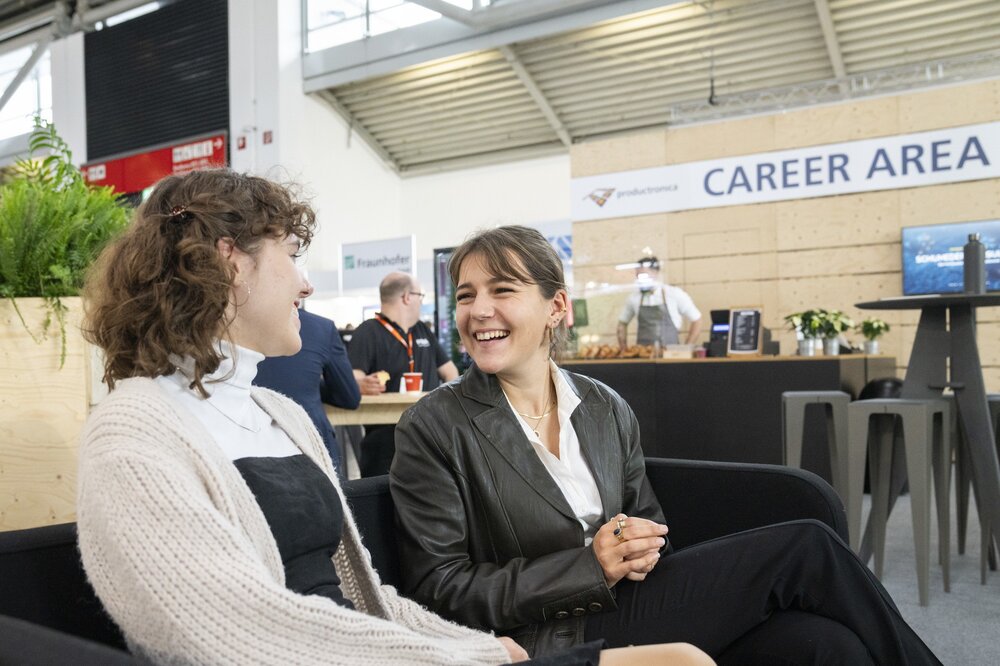 Two women are sitting on a black sofa in the catering area of the productronica CAREER AREA and laughing at each other.