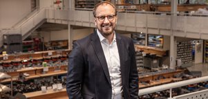 A man in a black suit with short hair and glasses leans smiling against the railing of a two-storey factory building.