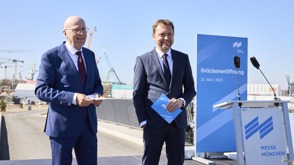 Zwei Männer in Anzügen lächeln bei der Eröffnungszeremonie einer Brücke, im Hintergrund sind ein blaues Schild und Kräne zu sehen.