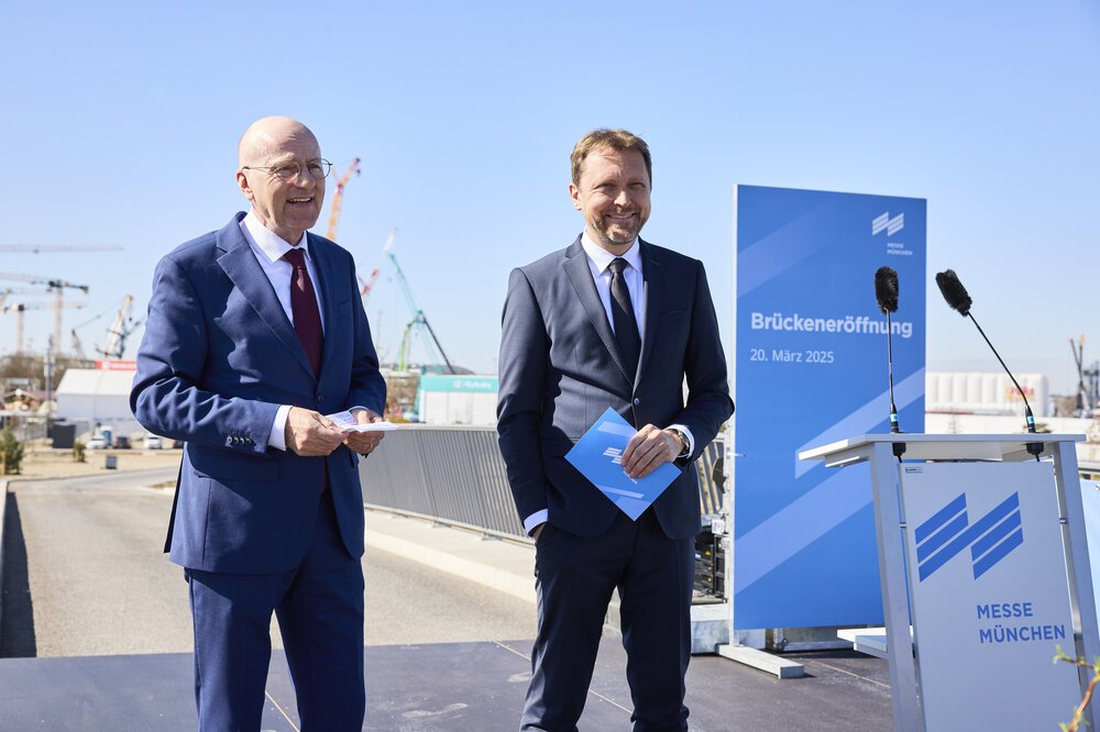 Zwei Männer in Anzügen lächeln bei der Eröffnungszeremonie einer Brücke, im Hintergrund sind ein blaues Schild und Kräne zu sehen.