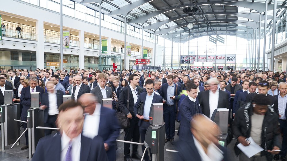 A large crowd of people in business attire passes through the turnstiles in a modern, spacious event hall.