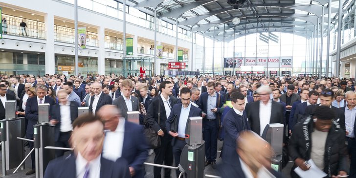 A large crowd of people in business attire passes through the turnstiles in a modern, spacious event hall.