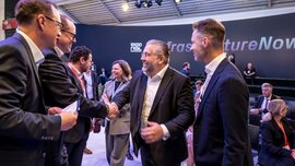 Business professionals greeting each other at EXPO REAL 2026 in Munich in front of the InfrastructureNow stage, surrounded by other trade fair participants.