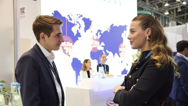 Two smiling people stand opposite each other in front of an exhibition stand. A counter with two women and an illuminated blue world map can be seen in the background.