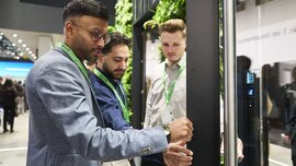 Three men in business attire with green lanyards around their necks inspect a metal door frame at an exhibition stand.