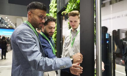 Drei Männer in Businesskleidung, mit grünen Schlüsselbändern um den Hals begutachten einen metallenen Türrahmen an einem Messestand.
