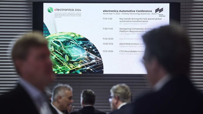 Blurred group of people in the foreground, behind them a display hangs on the wall showing the schedule of the electronica Automotive Conference.