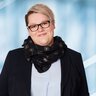 A woman with short blonde hair, glasses and a scarf as well as a blazer and blouse smiles.