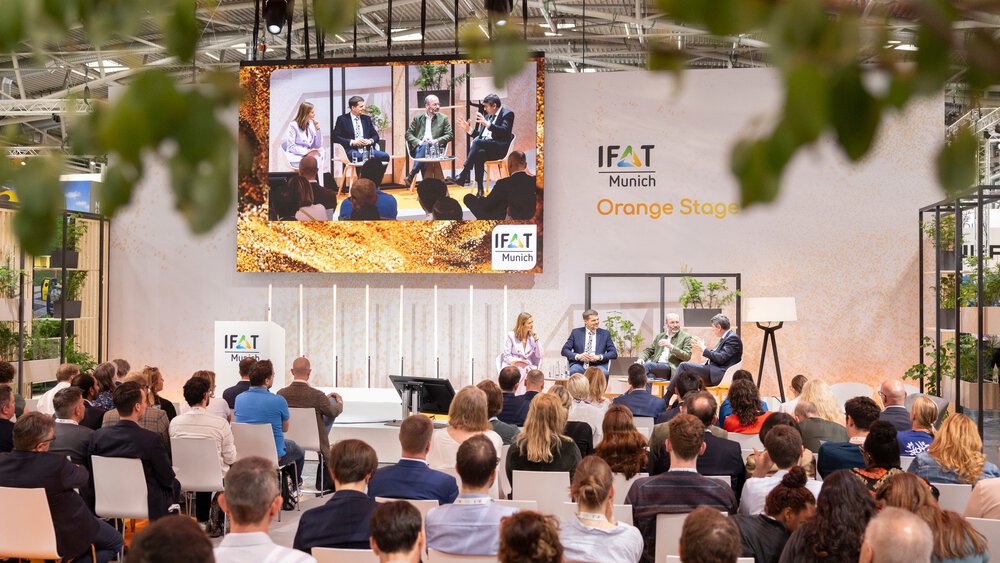 Audience seated in front of the “IFAT Munich – Orange Stage,” where a panel of four speakers is holding a discussion; a large screen above the stage displays a live feed of the talk, framed by plants and the trade fair setting.