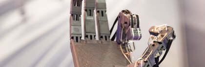Nahaufnahme einer metallischen Roboterhand mit angewinkeltem Daumen und Zeigefinger, vor einem unscharfen Hintergrund.
