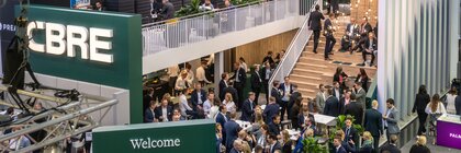 Überfüllter CBRE-Stand auf einer Wirtschaftsmesse mit Teilnehmern, die Kontakte knüpfen, und einer Treppe, die in eine obere Etage führt.