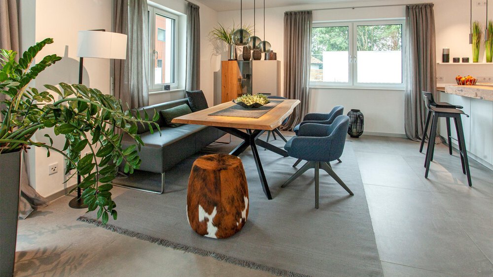 Modernes Wohnzimmer mit einem hölzernen Esstisch, grauen Stühlen, einer grünen Pflanze und natürlichem Licht durch ein Fenster.