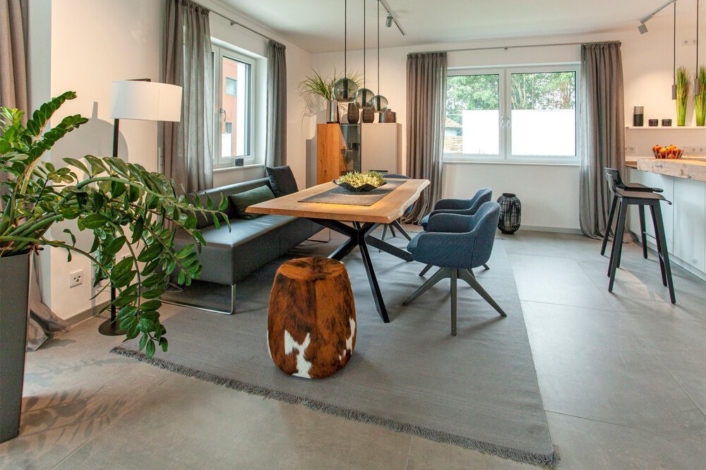 Modernes Wohnzimmer mit einem hölzernen Esstisch, grauen Stühlen, einer grünen Pflanze und natürlichem Licht durch ein Fenster.