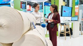 Zwei Frauen stehen an einem Stand auf der IFAT neben einem Stapel aus gerollten Stoffbahnen und unterhalten sich.
