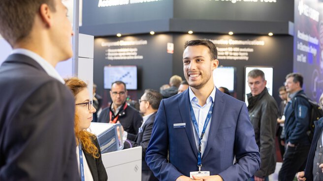 Ein Aussteller spricht lächelnd zu zwei Fachleuten im Vordergrund. Hinter ihm sind weitere Messestände und Besucher zu sehen.
