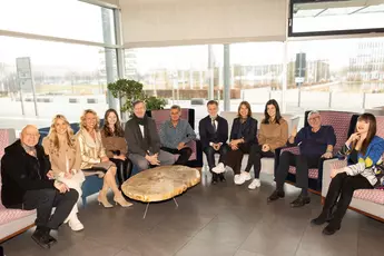 Eine Gruppenfoto von Menschen, die in einem hellen Raum zusammen sitzen, vor ihnen steht ein runder Holztisch.