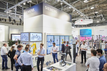 People at a Siemens booth with displays and robotic equipment in a large exhibition hall.