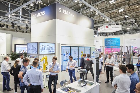 People at a Siemens booth with displays and robotic equipment in a large exhibition hall.
