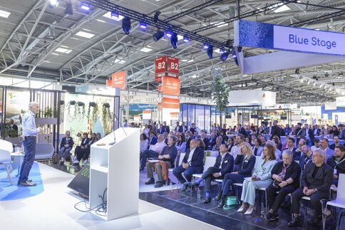 A man is standing on a stage, speaking to an audience. Information signs hang from the ceiling of the hall, including one labelled “Blue Stage”.