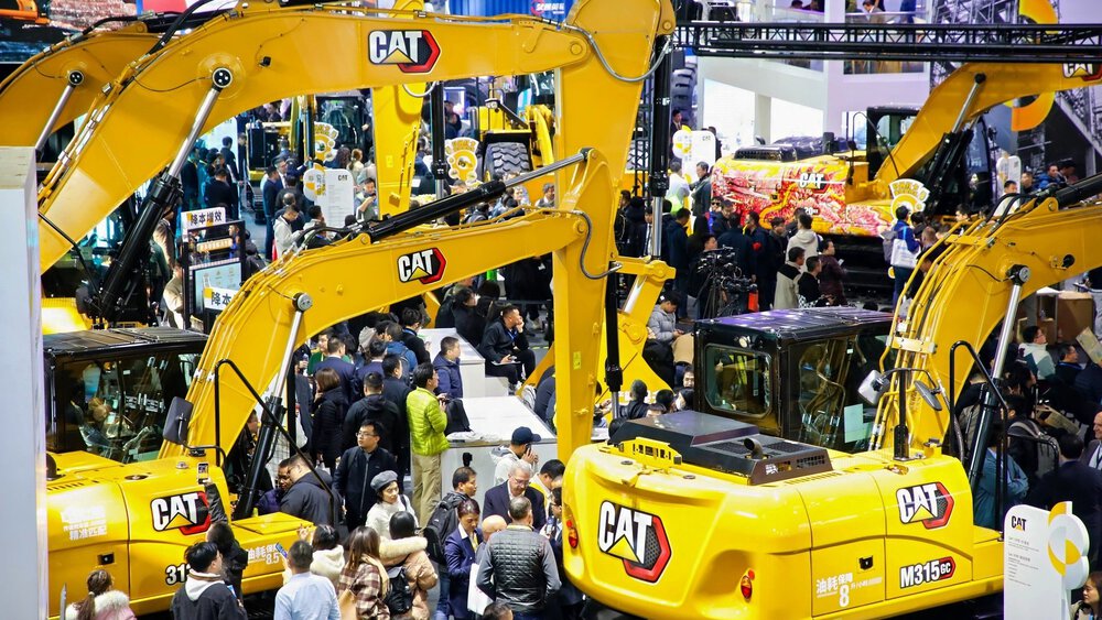 A crowd at a trade show featuring large yellow Caterpillar excavators and construction equipment.