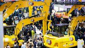 A crowd at a trade show featuring large yellow Caterpillar excavators and construction equipment.