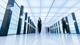 Eine schwarz gekleidete Frau geht durch einen futuristischen, hell erleuchteten Flur, der von hohen, beleuchteten Tafeln gesäumt ist.