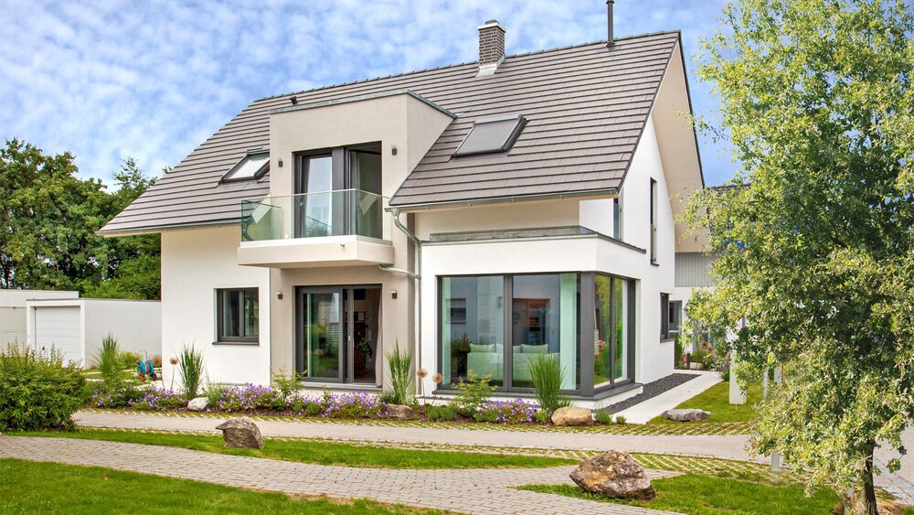Modernes zweistöckiges Haus mit großen Fenstern, einem Balkon und einem Satteldach, umgeben von einem angelegten Garten.