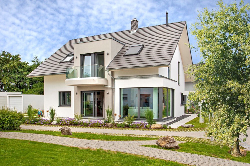 Modernes zweistöckiges Haus mit großen Fenstern, einem Balkon und einem Satteldach, umgeben von einem angelegten Garten.