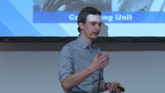 Ein Mensch spricht auf der Bühne und hält einen Clicker, im Hintergrund einen Projektionsbildschirm mit Text.