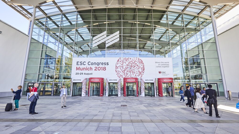 People are walking past a glass building with a banner hanging above its entrance that reads “ESC Congress Munich 2018, August 25–29.”