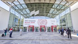 People are walking past a glass building with a banner hanging above its entrance that reads “ESC Congress Munich 2018, August 25–29.”